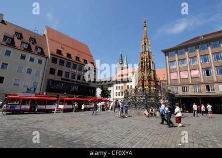 A red tourist train parked in Nuremberg's main market square, (Hauptmarkt), Bavaria, Germany next to the 14th-century Schoner Brunnen. The golden Stock Photo