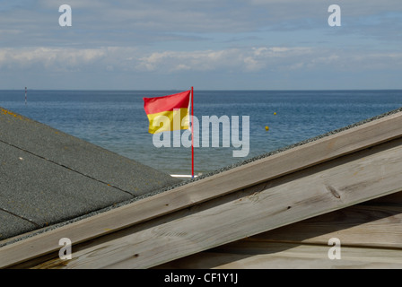 A bathing flag seen between the roofs of two beach huts. The bathing flag marks one end of a section of beach patrolled by lifeg Stock Photo