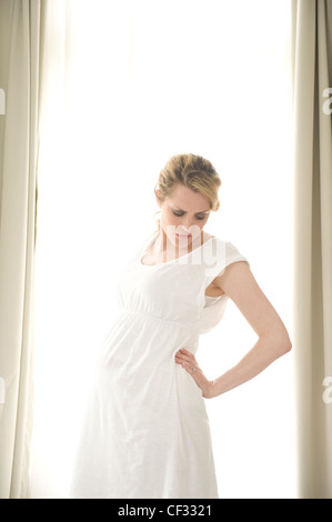 Pregnant female fair hair tied into ponytail, wearing a white dress, standing by the window hand touching back, unsmiling, Stock Photo