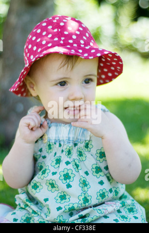 A female baby fair hair, wearing a pink polka dot summer hat and green floral summer dress, finger in mouth, unsmiling, Stock Photo