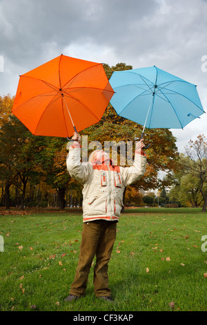 Boy in autumn park. Holds over head two colour umbrellas under cloudy sky. Stock Photo