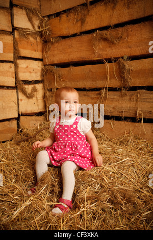 Little girl is sitting on pile of straw in hayloft against the wall of boards. Vertical format. Stock Photo