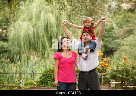 Little girl in red dress together with parents in summer garden. Girl sits on shoulders at father. Stock Photo