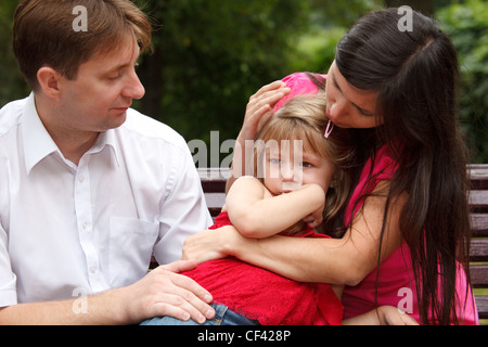 Parents calm crying girl on walk in summer garden. Mum embraces daughter. Stock Photo