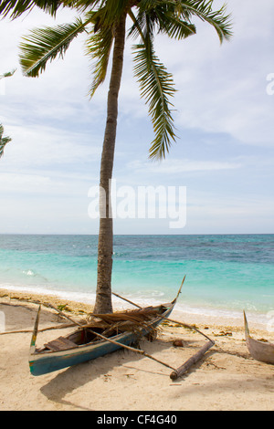 A small boat rests under a shady palm tree on a remote island beach Stock Photo