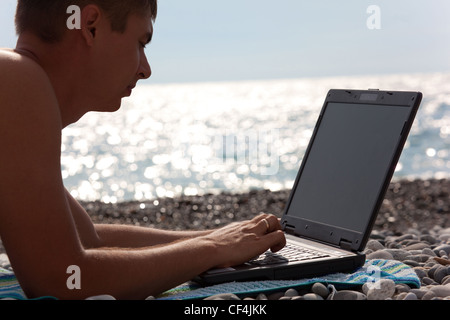 young man working on his notebook on beach. close-up portrait of guy with laptop lying on seacoast. Stock Photo