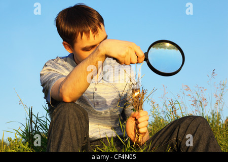 one man wearing shirt and jeans with magnifier is sitting on a meadow and burning grasses Stock Photo