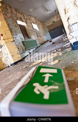 Emergency exit sign on the floor of a derelict hospital, with arrow pointing toward corridor with peeling walls. Stock Photo