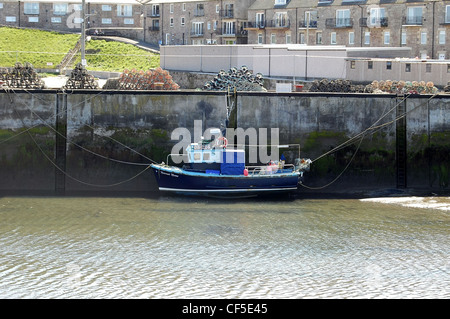 Seahouse harbour at low tide with boat resting on the muddy seabed with fishing gear stacked on top of harbour wall Stock Photo