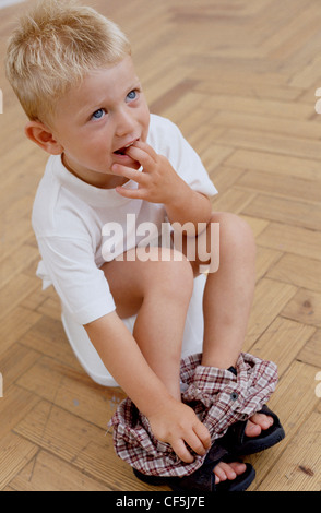 Male toddler blonde hair wearing a white t shirt, checkered trousers pulled down and sandals, sitting on potty, fingers in Stock Photo