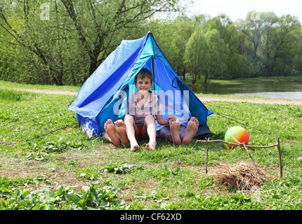 From blue tents on a grassy glade meadow legs stick out and a little boy sitting in it and smile Stock Photo