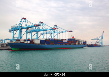 Big cargo boat and small barge docked to industrial port with blue cranes Stock Photo