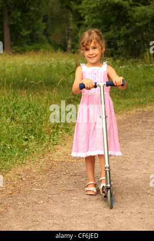 beautiful little girl in pink dress rides scooter on nature. forest could be seen in background Stock Photo