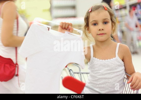 little girl sitting in store cart and holding hanger with white shirt, looking at camera Stock Photo