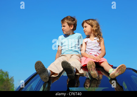 little boy and girl sitting on car roof on blue sky, looking to left side Stock Photo