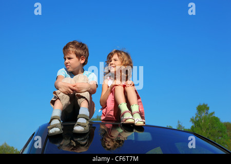 little boy and girl sitting on car roof on blue sky, hands on legs Stock Photo