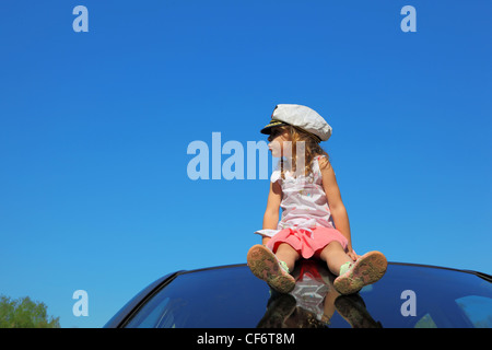 little girl with captain cap sitting on car roof watching on left side, blue sky Stock Photo