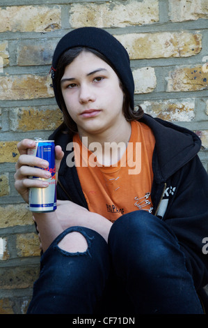 Teenage boy wearing black beanie orange t shirt and black hooded top and black jeans sitting on wall knees up holding can of Stock Photo