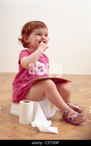 Female toddler ginger hair wearing a short pink dress and sandals, sitting on potty, holding a book, finger in mouth, toilet Stock Photo