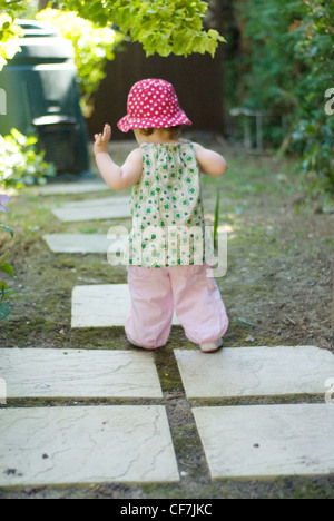 A female baby, wearing a pink polka dot summer hat, green floral dress and pink trousers, walking in the garden Joel Penn Stock Photo