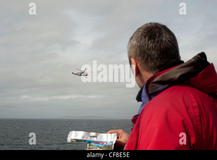 A man in red jacket watches an RAF Sea King rescue helicopter hover close by above the sea in Scotland.. Stock Photo