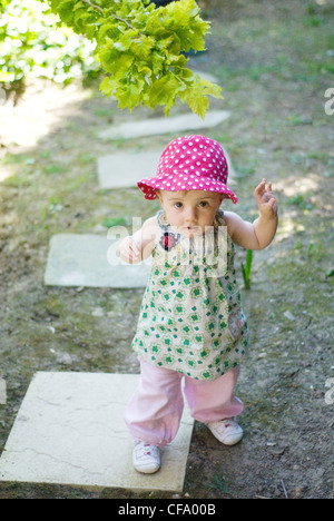 A female baby, wearing a pink polka dot summer hat, green floral top, white trainers and pink trousers, unsmiling, looking up Stock Photo