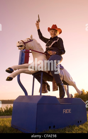 Senior woman riding mechanical horse in cowboy costume, Canada. Stock Photo