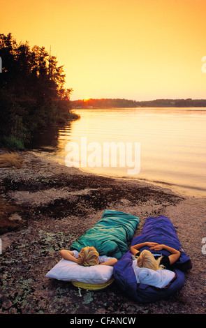 Two children in sleeping bags along shoreline of lake, Whiteshell Provincial Park, Manitoba, Canada Stock Photo