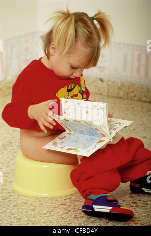 Female child with blonde hair in bunches sitting on potty reading book Stock Photo