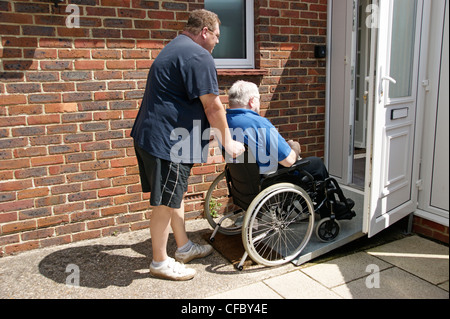 male carer / son pushing elderly man in wheelchair on ramp access to property Stock Photo