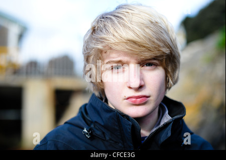 Angry blond-haired white teenage boy  in a dark jacket stands outside and stares directly at the camera Stock Photo
