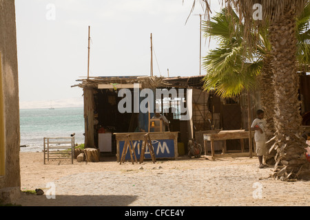 Sal Rei Boa Vista Cape VerdeLocal man building a chair in a water front carpenters' shack young boy playing around palm tree Stock Photo