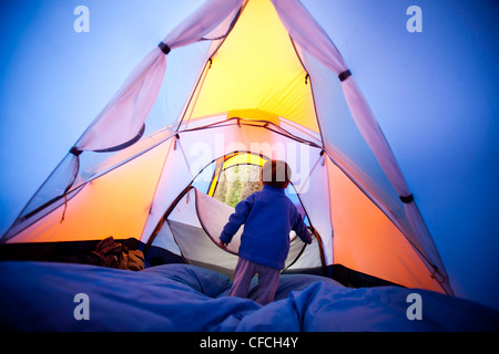 a little boy opens and closes the zippered tent door while he stand on a blue sleeping bag. The tent is orange and blue. The cam Stock Photo