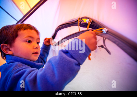 a little boy opens and closes the zippered tent door while he stands on a blue sleeping bag. The tent is orange and blue. The ca Stock Photo