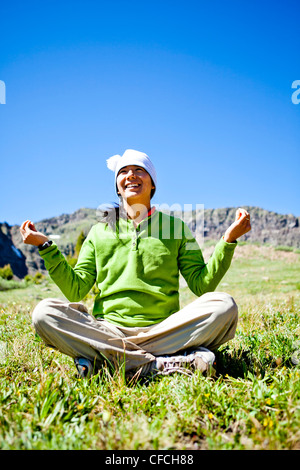 a woman smiles as she sits cross legged in an alpine meadow with wildflowers doing a Buddha pose. They are just off the Continen Stock Photo