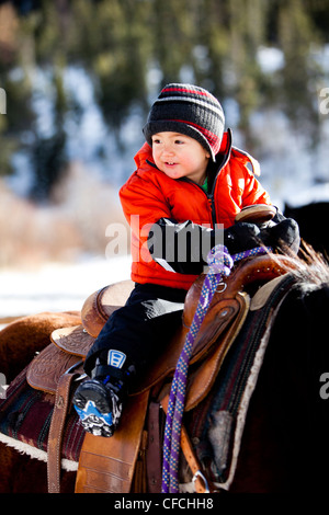 a little boy rides a horse while bundled up in a warm jacket and stocking cap / hat on a cold winter day with snow on the ground
