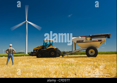 A man in harvested barley field with tractor and grain wagon, viewing wind turbines, near St. Leon, Manitoba, Canada