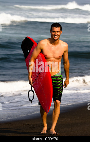 A male surfer wearing board shorts walks up the beach at Pasquales, Mexico while holding a red surfboard under his arm.