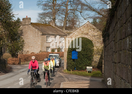 Group of cyclists travelling on bikes, cycling & pedalling on country road through scenic rural village - Bolton Abbey, North Yorkshire, England, UK Stock Photo