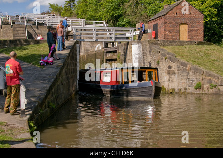 Sunny boat trip (people stand, watching as narrowboat steers out of bottom lock chamber) - Five Rise Locks, Bingley, Leeds Liverpool Canal, England. Stock Photo