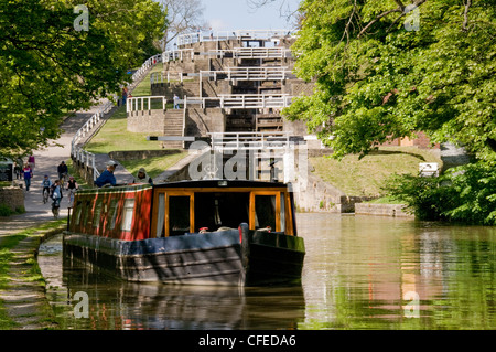Narrowboat travelling on scenic rural waterway (Five Rise Locks, Leeds Liverpool Canal) with man & woman aboard - Bingley, West Yorkshire, England, UK Stock Photo