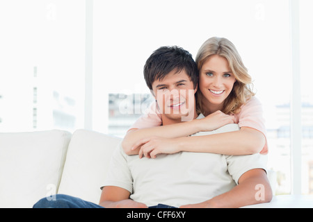 Man sitting on the couch with woman holding him from behind as they smile Stock Photo