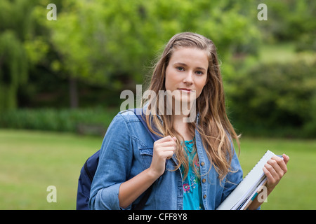 Young woman standing up in a park while holding a notebook and a backpack Stock Photo