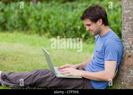 Young smiling man leaning against a tree while using his laptop Stock Photo