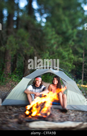 A young adult couple laugh and smile sitting in their tent next to a campfire in Idaho. Stock Photo