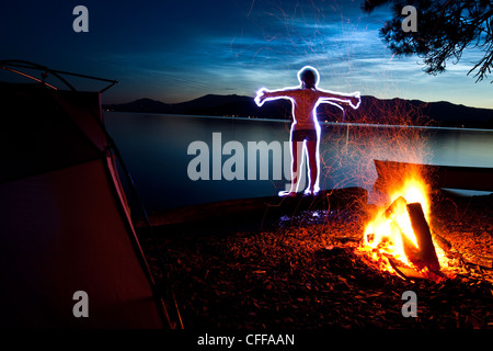 A light painting of a woman doing yoga next to her tent and campfire on a camping trip along the shores of a lake in Idaho. Stock Photo