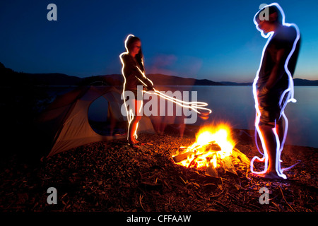 A light painting of a adult couple standing next to their tent and campfire on a camping trip along the shores of a lake in Idah Stock Photo