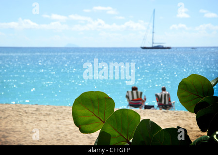 Caribbean beach scenery with couple relaxing in chairs on the beach. In the horizon a sailboat is anchoring out. In the foreground leaves of seagrapes. Stock Photo