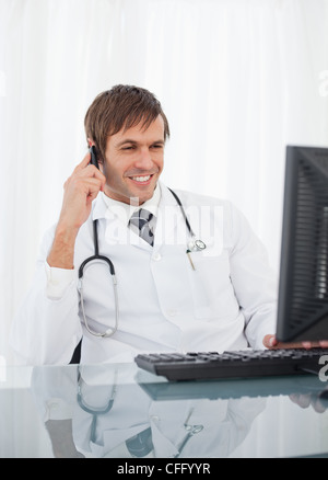 Smiling doctor looking at his computer while using his mobile phone Stock Photo