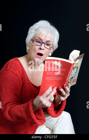 Attractive senior lady in bright clothes with a surprised look on face sitting reading book taken against a black background Stock Photo